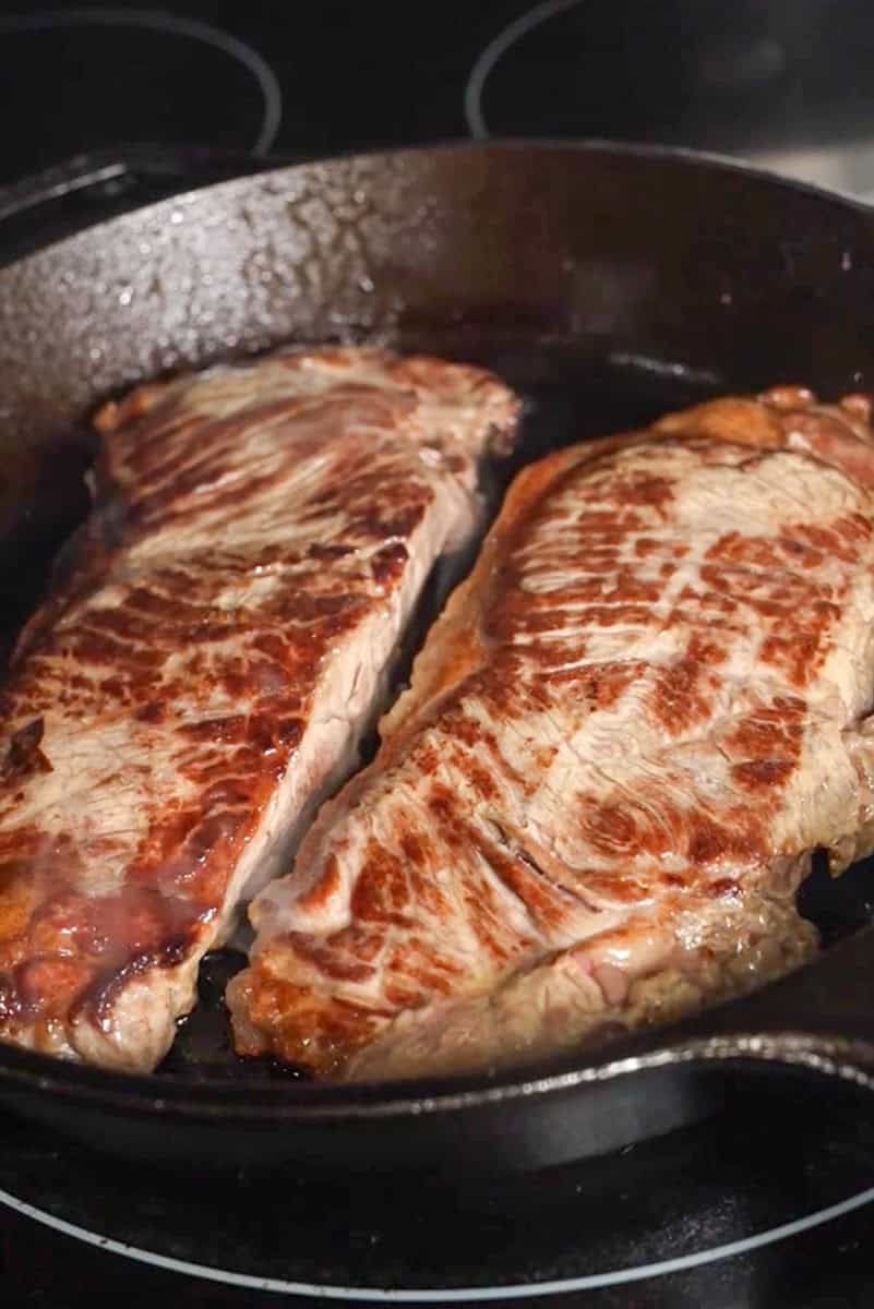 Two New York strip steaks searing in a cast-iron skillet