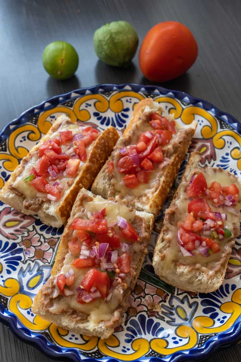 Esta Molletes Mexicanos Receta es un plato que incorpora frijoles refritos, queso blanco derretido, tomate, cebolla morada y limón verde.