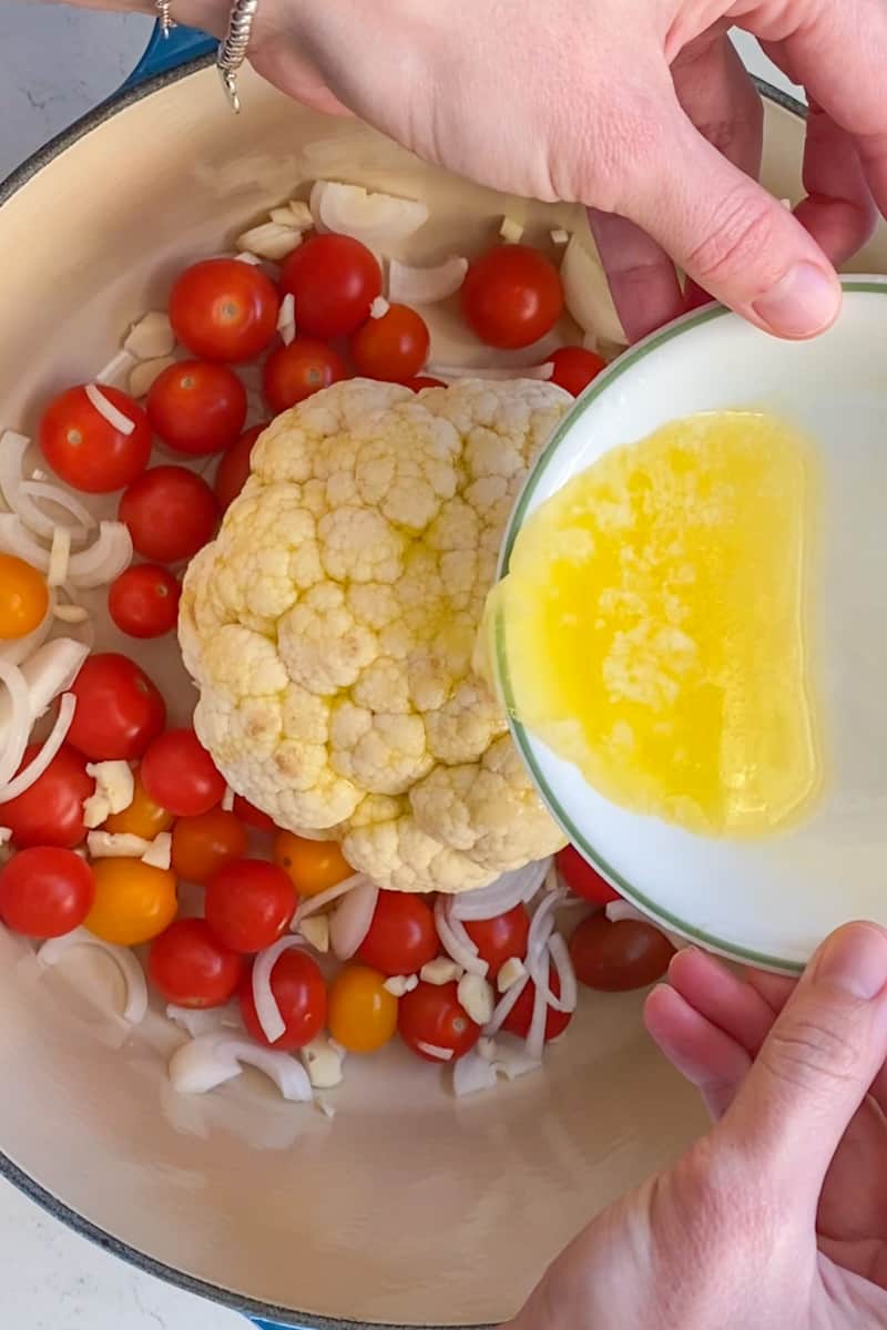 Place the cauliflower in a Dutch oven. Drizzle olive oil over the cauliflower and use your hands to evenly coat. Place the cauliflower, core side down. Add cherry tomatoes, onion, and garlic around the cauliflower. Pour white wine onto the cauliflower as well as butter.