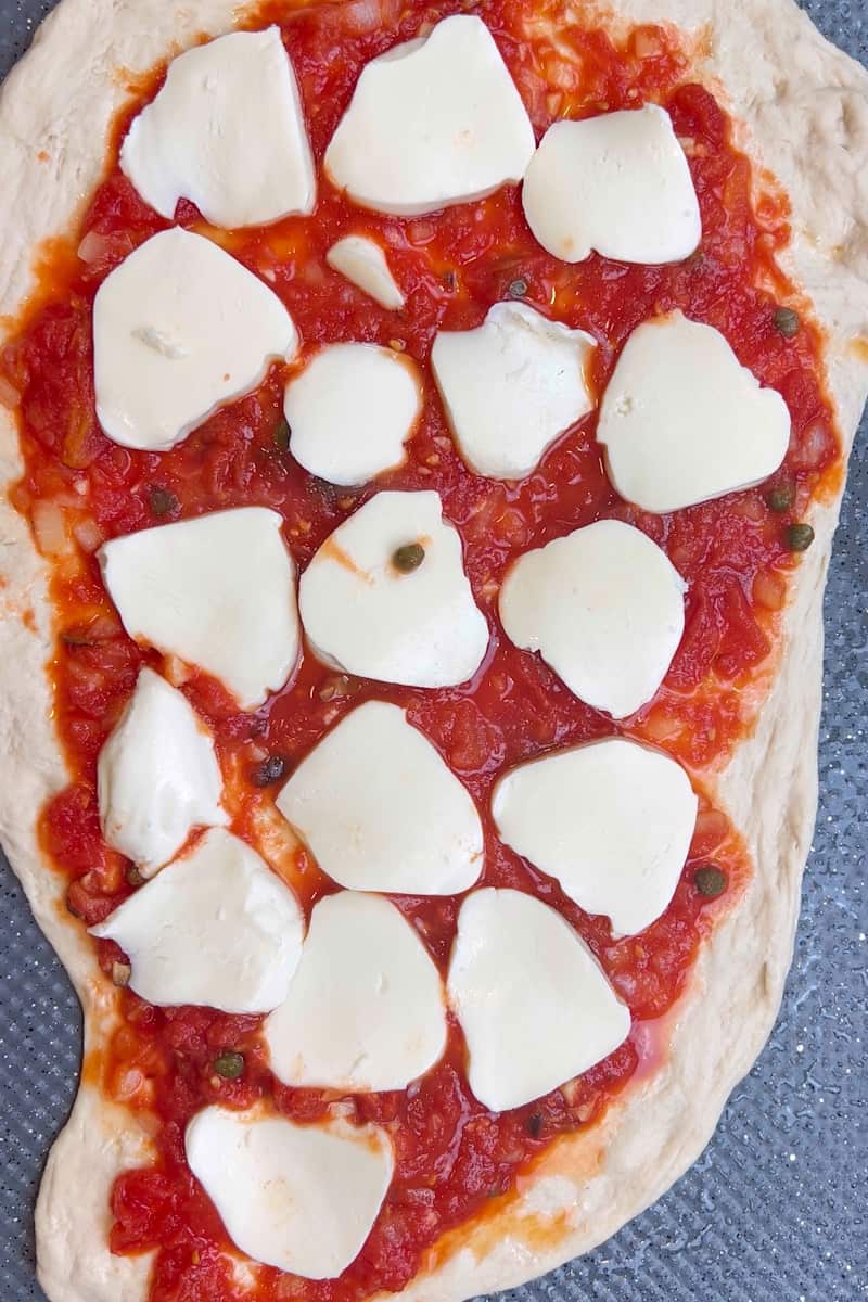 With a spoon, add the puttanesca sauce on the pizza. Slice the mozzarella and add in an even layer.